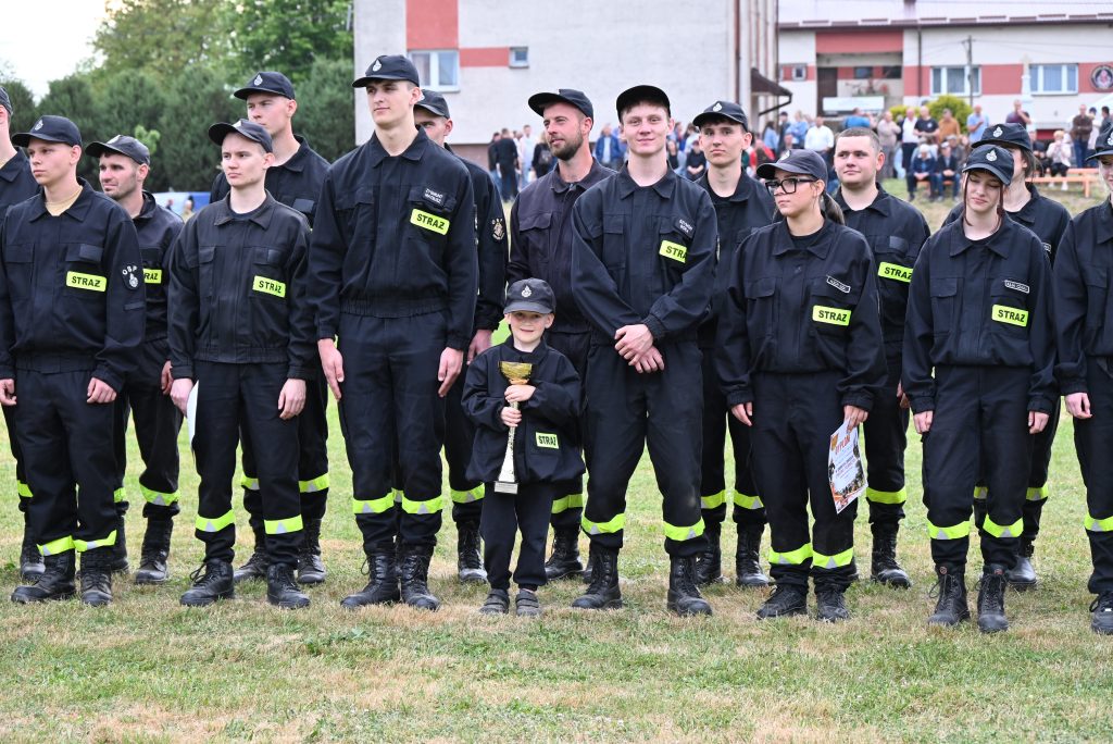 Grupa młodych strażaków ubranych w czarne mundury z żółtymi oznaczeniami „STRAŻ”, stojących na trawie, jeden z nich trzyma złoty puchar.
