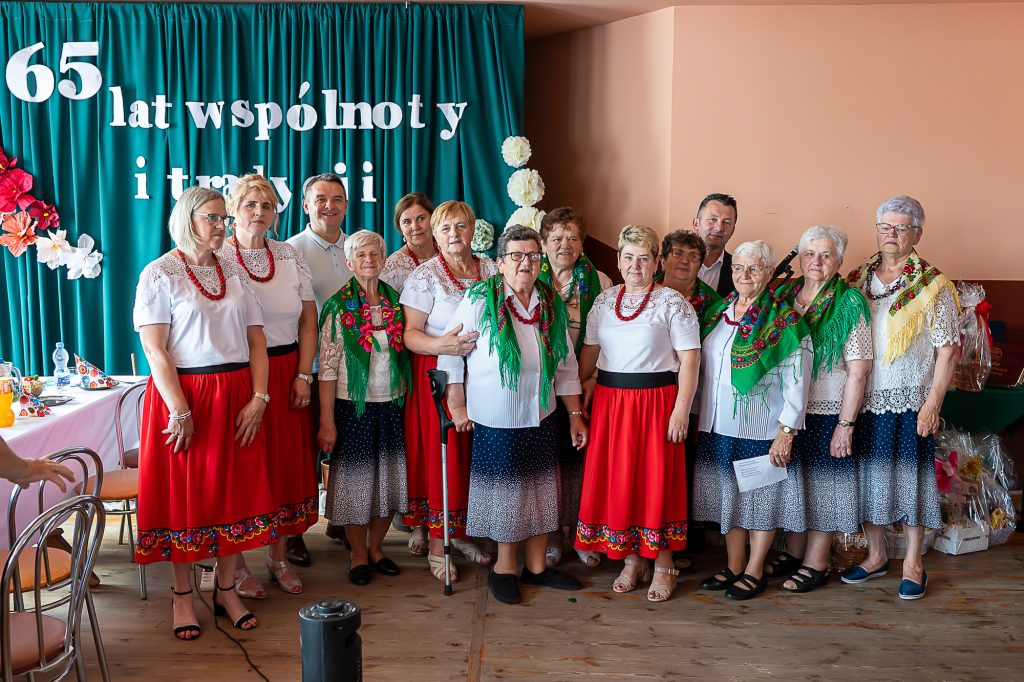 Grupa osób w tradycyjnych strojach na tle zielonej kurtyny z białymi literami i dekoracjami kwiatowymi, drewniana podłoga, jasne wnętrze.