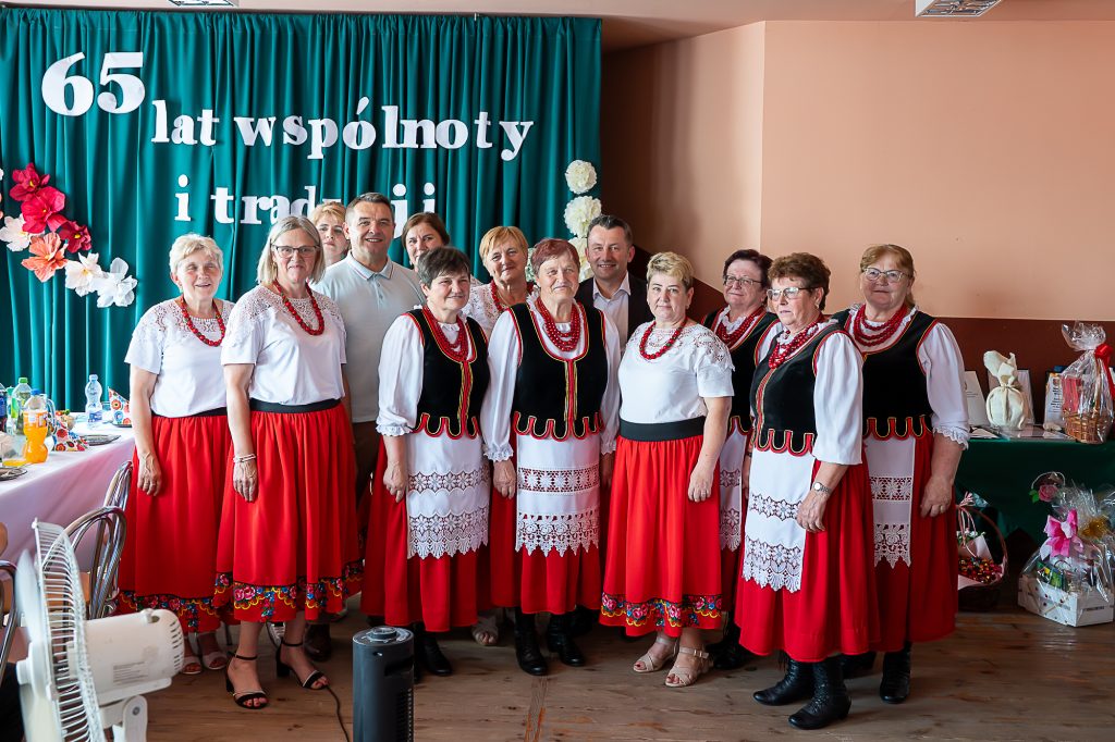 Grupa osób w tradycyjnych strojach z czerwonymi spódnicami i koralami na tle zielonej kurtyny z napisem "65 lat wspólnoty i tradycji" oraz dekoracjami kwiatowymi.