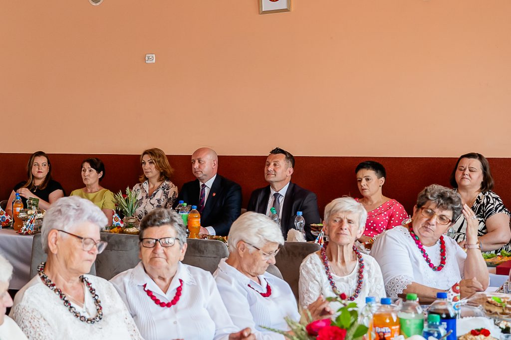 Grupa osób siedzących przy stole z napojami i przekąskami, ściana w tle w dwóch odcieniach pomarańczowego, na stole kolorowe serwetki i kwiaty.