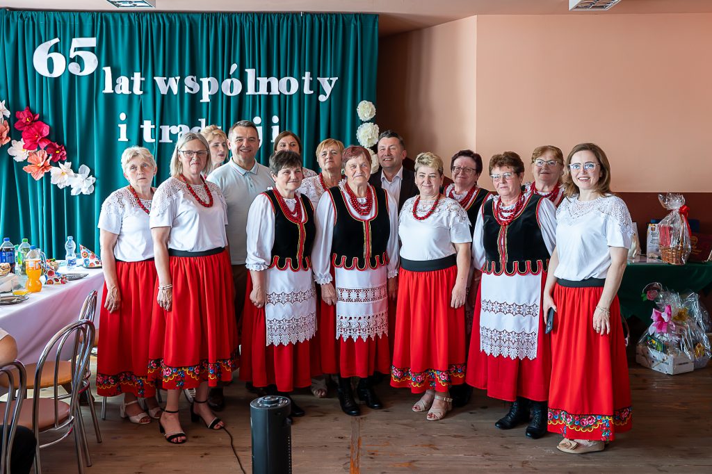 Grupa 14 osób, kobiety w białych bluzkach z koronką i czerwonych spódnicach, mężczyźni w jasnych koszulach, stoją na tle zielonej zasłony z napisem.