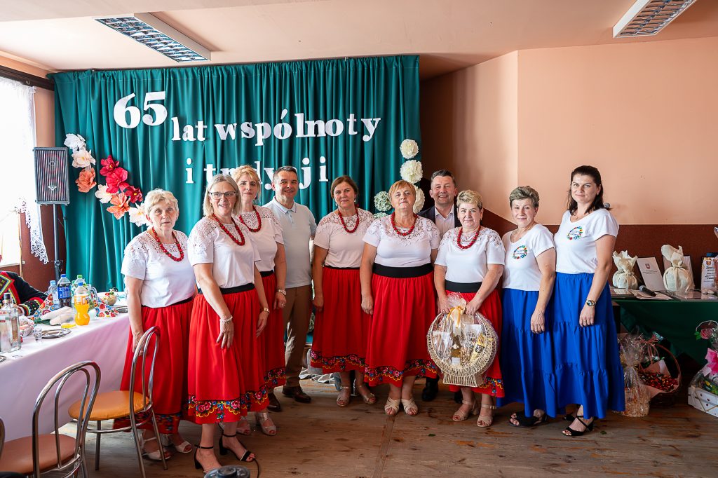 Grupa dziesięciu osób w tradycyjnych strojach, kobiety w białych bluzkach i czerwonych lub niebieskich spódnicach, mężczyzna w jasnej koszulce, na tle zielonej kurtyny z napisem.