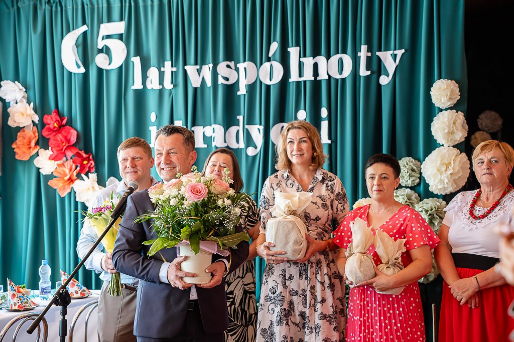 Grupa uśmiechniętych osób trzymających kwiaty i prezenty na tle zielonych zasłon z białymi literami oraz dekoracjami z dużych papierowych kwiatów.