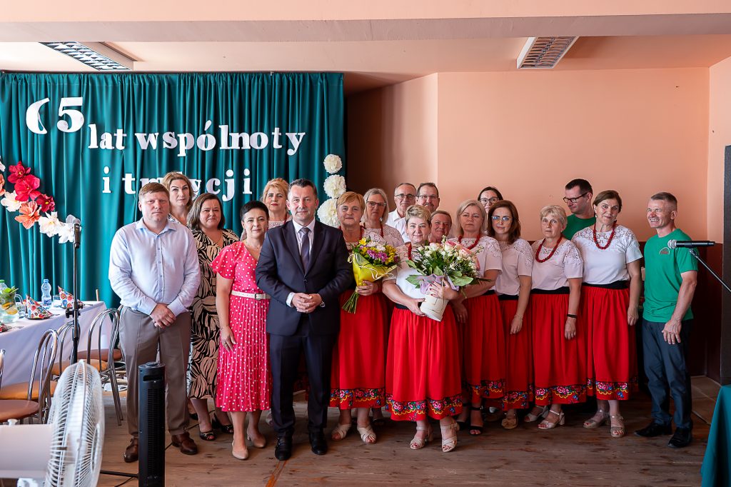 Grupa ludzi na tle zielonych zasłon z białym napisem, kobiety w białych bluzkach i czerwonych spódnicach, mężczyźni w koszulach i garniturze, dwie kobiety trzymają bukiety kwiatów.