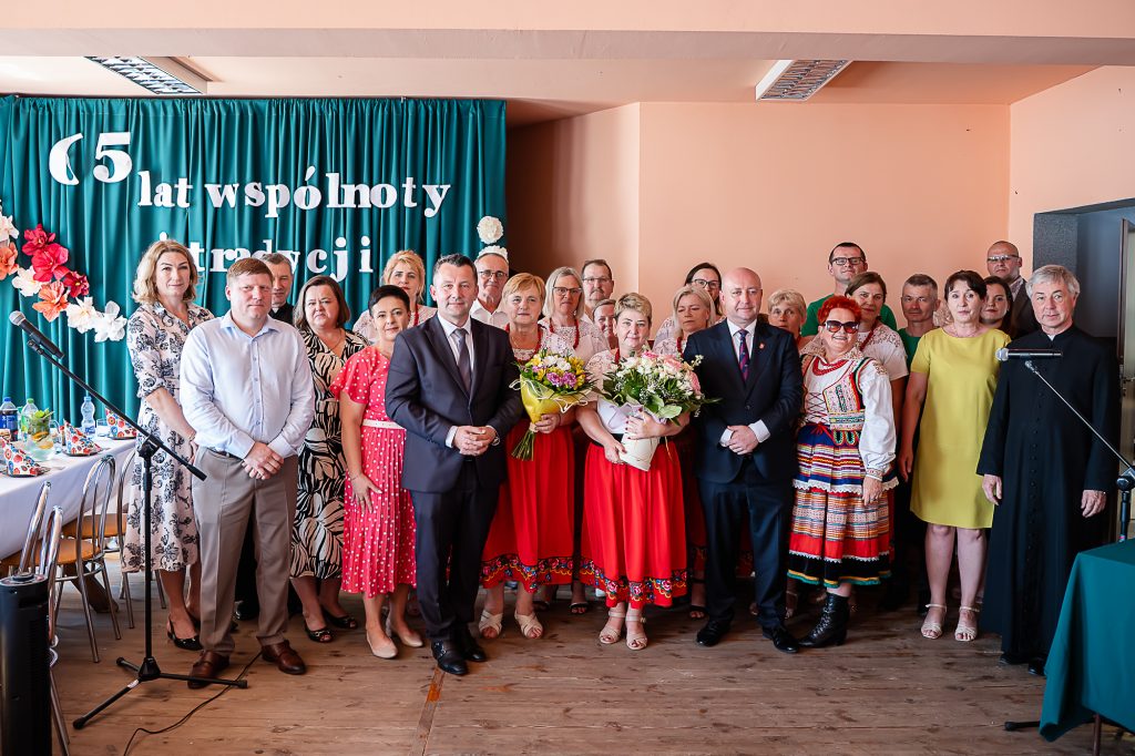 Grupa osób w różnorodnych strojach, w tym tradycyjnych, stojąca na tle zielonej zasłony z białym napisem, z kwiatami i mikrofonami po bokach.