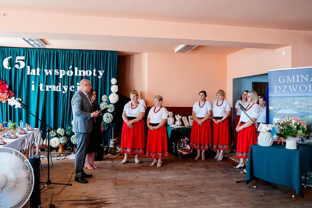 Grupa kobiet w białych bluzkach i czerwonych spódnicach stoi w sali z drewnianą podłogą, obok stoi mężczyzna w garniturze, za nimi niebieska kurtyna z białymi literami.