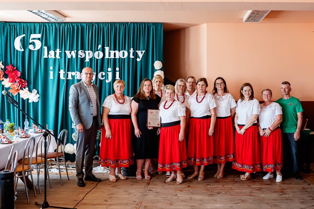 Grupa osób na tle turkusowej zasłony z napisem "65 lat wspólnoty i tradycji", kobiety w białych bluzkach i czerwonych spódnicach, mężczyźni w garniturze i zielonej koszulce.