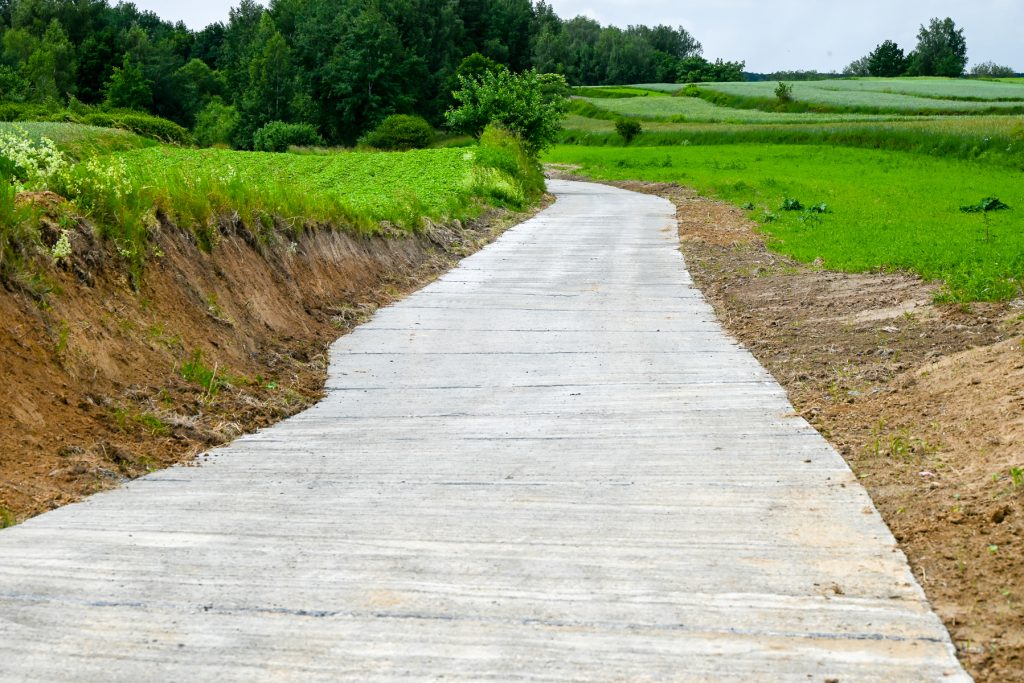 Betonowa droga na tle zielonych pól i drzew, po bokach widoczna ziemia i niska roślinność.