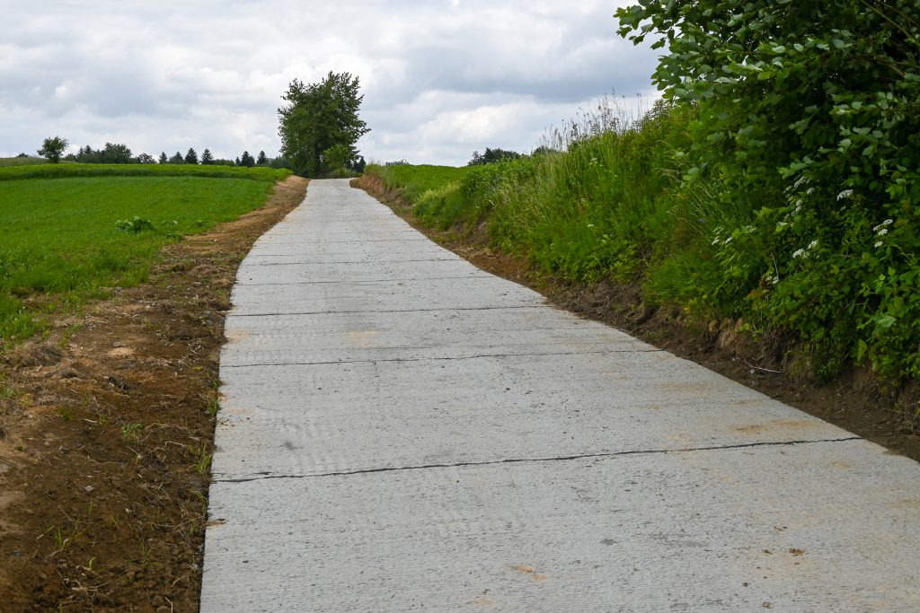 Wąska betonowa droga na tle zielonych pól i krzewów, pod pochmurnym niebem.
