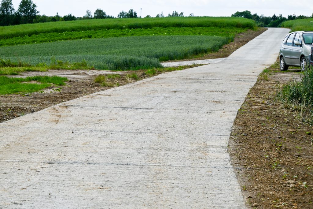 Szara betonowa droga na tle zielonych pól i drzew, po prawej stronie dwa zaparkowane srebrne samochody, przydrożna ziemia z trawą.