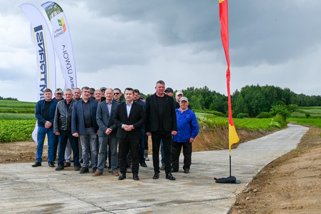 Grupa mężczyzn w różnym wieku stoi na betonowej drodze na tle zielonych pól i drzewa, nad nimi pochmurne niebo, obok flagi z logo i napisem.