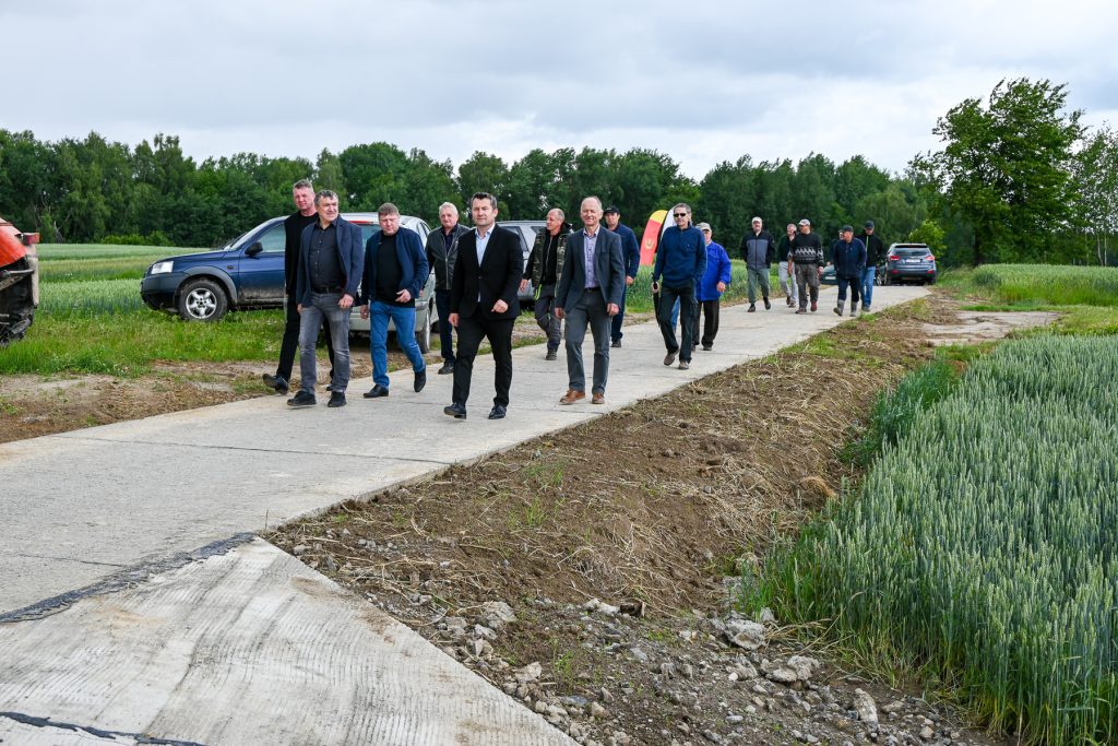 Grupa mężczyzn w różnym wieku idąca po betonowej drodze obok zielonych pól i lasu pod pochmurnym niebem.
