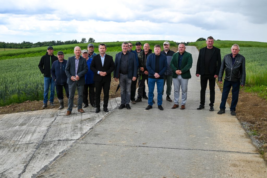 Grupa mężczyzn w różnym wieku stojących na betonowej drodze otoczonej zielonymi polami pod pochmurnym niebem.