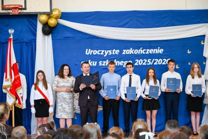 Grupa osób w formalnym ubraniu na tle niebieskiego baneru z białym napisem i białą dekoracją, trzymających niebieskie teczki, obok flagi Polski.