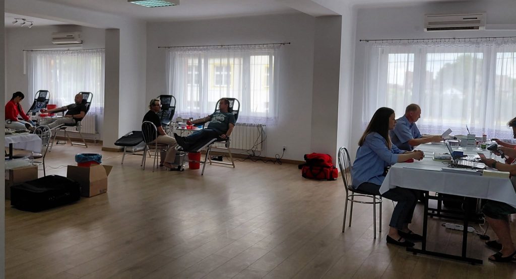 Wnętrze jasnego pomieszczenia z ludźmi na krzesłach, białe ściany, drewniana podłoga, duże okna z białymi firankami, stolik z laptopami i sprzętem medycznym.