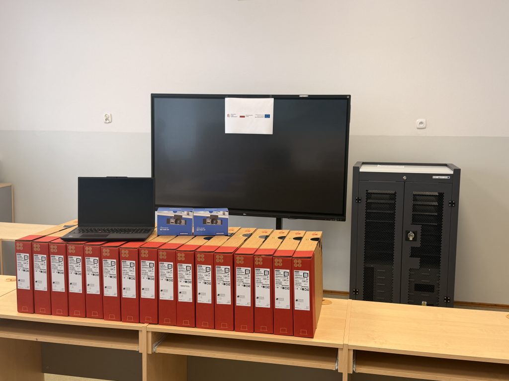 Czarny laptop na stole obok dwóch niebieskich pudełek i rzędu czerwonych segregatorów, za nimi duży czarny monitor z kartką oraz szafa serwerowa.