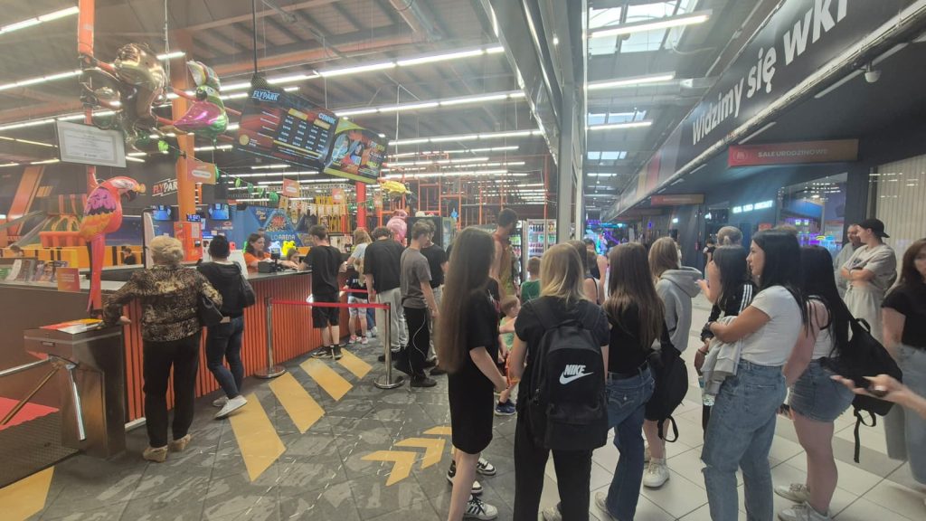 Wnętrze centrum rozrywki z kolejką ludzi, kolorowe balony w kształcie ptaków, jasne oświetlenie, tablice z cenami, osoby w różnym wieku, podłoga z żółtymi oznaczeniami.