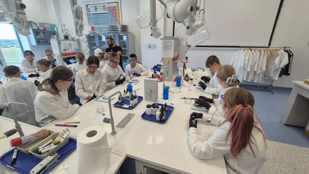 Laboratorium z białymi blatami, kilkanaście osób w białych fartuchach i okularach ochronnych, na stole szkło laboratoryjne, dozowniki i chemikalia, szafka z laborkami w tle.
