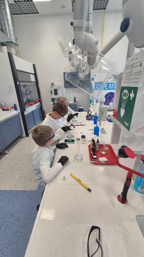 Laboratoryjne stanowisko z trzema osobami w białych fartuchach i czarnych rękawiczkach, szkło laboratoryjne, urządzenia, niebieskie i białe wnętrze.
