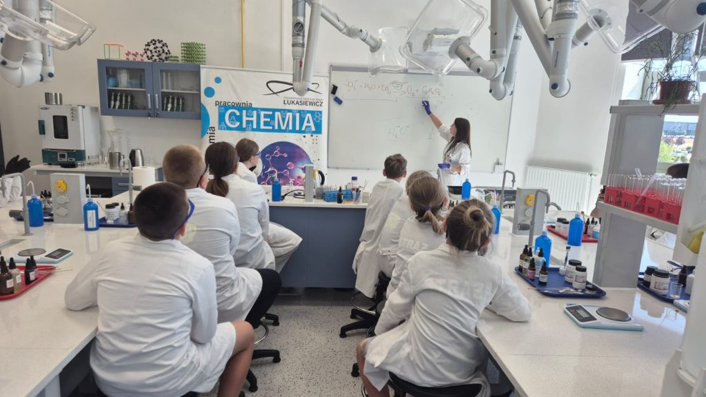 Laboratorium chemiczne z uczniami w białych fartuchach siedzącymi przy stołach, nauczycielka pisze wzory chemiczne na białej tablicy, na stole sprzęt laboratoryjny i butelki.
