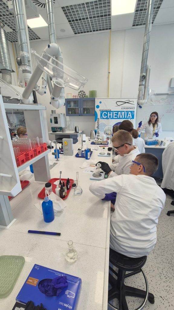 Laboratorium chemiczne z uczniami w białych fartuchach i okularach ochronnych, sprzęt laboratoryjny, probówki, pipety, chemikalia na stole, tablica z napisem "CHEMIA".