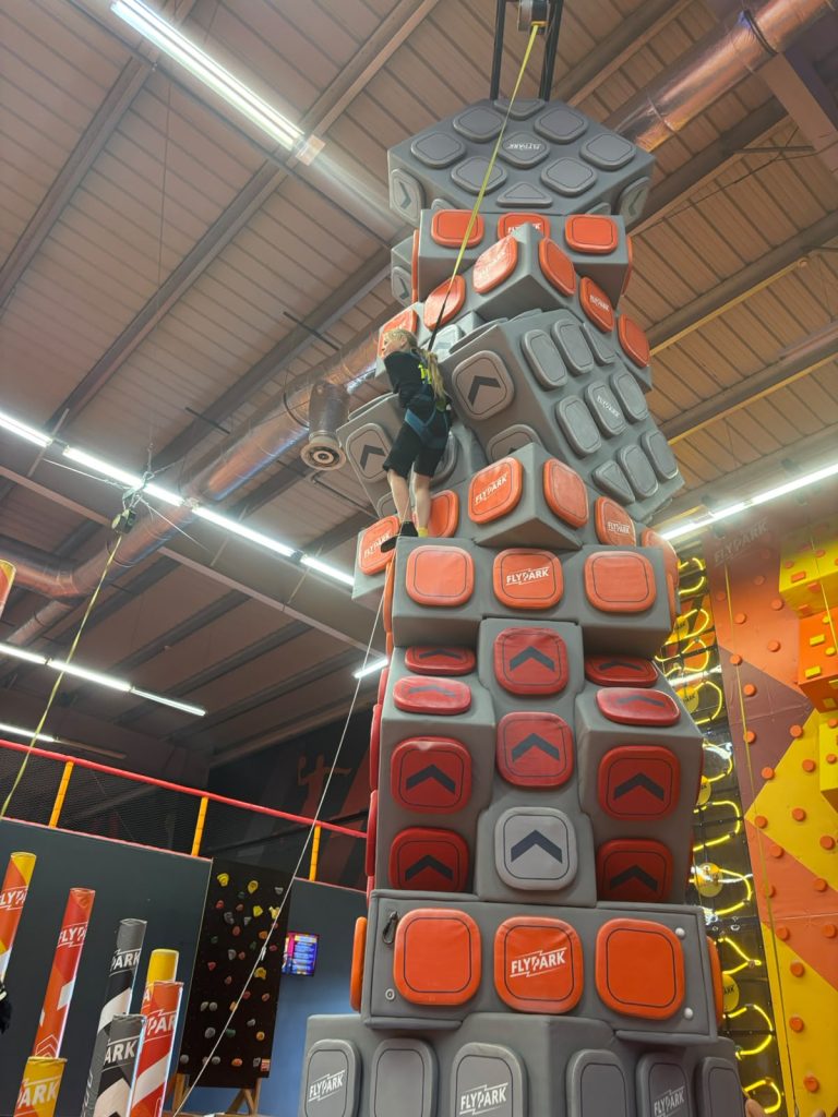 Wysoka, wielokolorowa ścianka wspinaczkowa z szarymi, czerwonymi i pomarańczowymi wypustkami oraz napisem "FlyPark", a na niej wspinająca się osoba w uprzęży.
