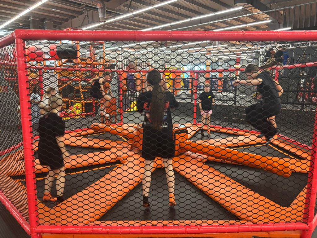 Cztery osoby skaczą na pomarańczowo-czarnych trampolinach otoczonych czerwonym stelażem z czarną siatką, w tle konstrukcje i oświetlenie hali sportowej.