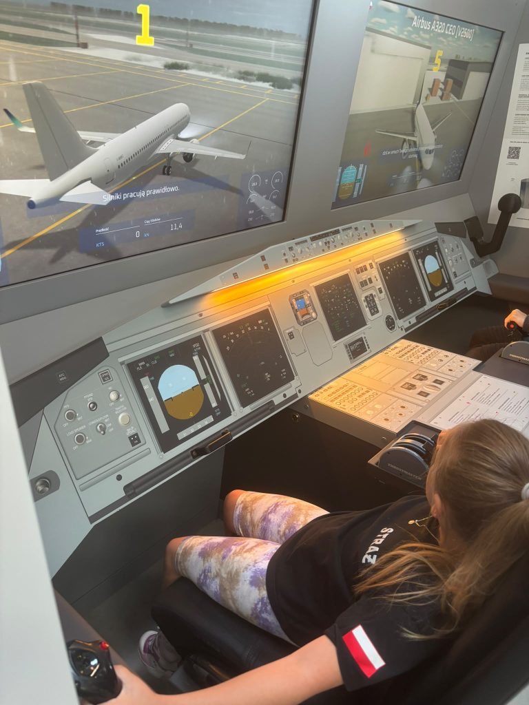 Kokpit symulatora lotu z dwoma ekranami pokazującymi samolot Airbus A320, panelami kontrolnymi i osobą siedzącą na fotelu pilota.