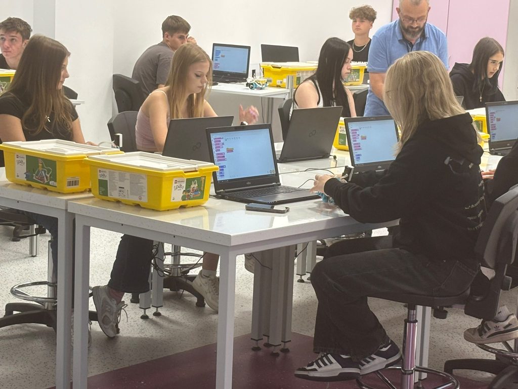 Grupa młodych osób siedzi przy białych stołach z laptopami i żółtymi plastikowymi pudełkami, w jasnym pomieszczeniu, na obrotowych krzesłach.