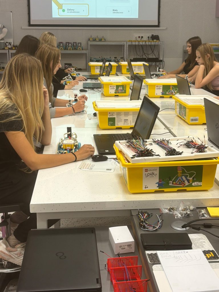 Grupa młodych osób siedzi przy białym stole z laptopami, żółtymi pudełkami LEGO Education Spike i robotami, w tle projektor wyświetlający fragment ekranu.
