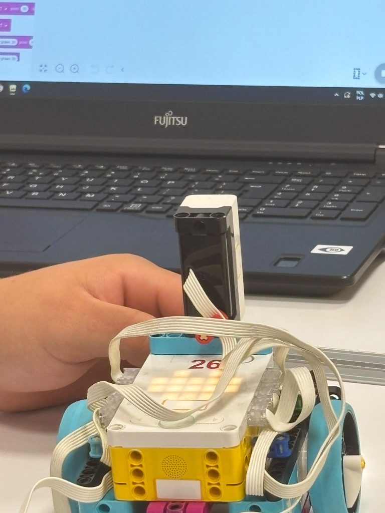 Dłoń trzymająca czarno-biały czujnik podłączony do kolorowego robota z kablami, na tle otwartego laptopa Fujitsu z niebieskim ekranem.