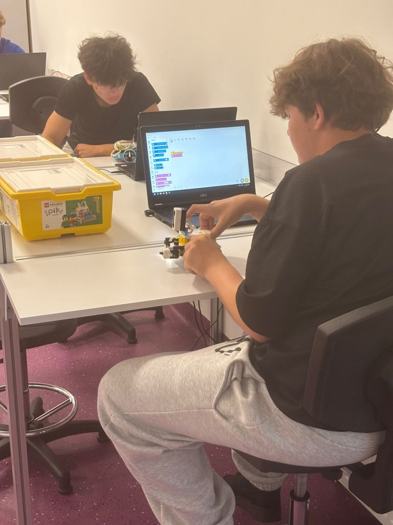 Dwóch chłopców siedzi przy białych stołach, jeden manipuluje małym robotem, na stole laptop z otwartym programem do kodowania, obok żółta skrzynka LEGO Education Spike.