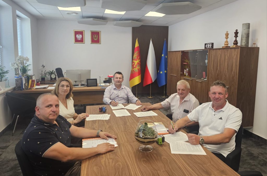 Cztery osoby siedzące przy drewnianym stole z dokumentami, za nimi trzy flagi i drewniana szafa z trofeami, jasne ściany i sufit z panelami akustycznymi.