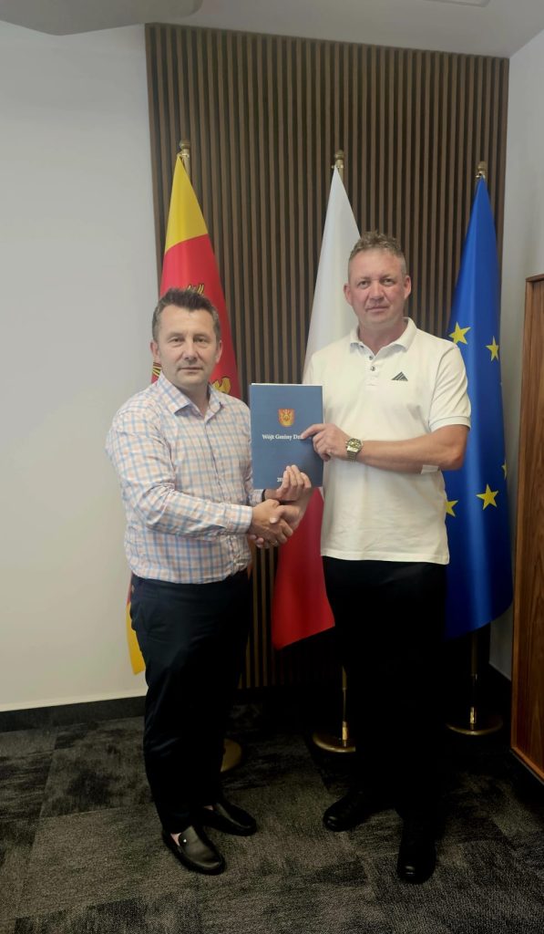 Dwaj mężczyźni ściskają sobie dłonie, jeden trzyma niebieski dokument, w tle trzy flagi: żółto-czerwono-czarna, biała i niebieska z żółtymi gwiazdami.