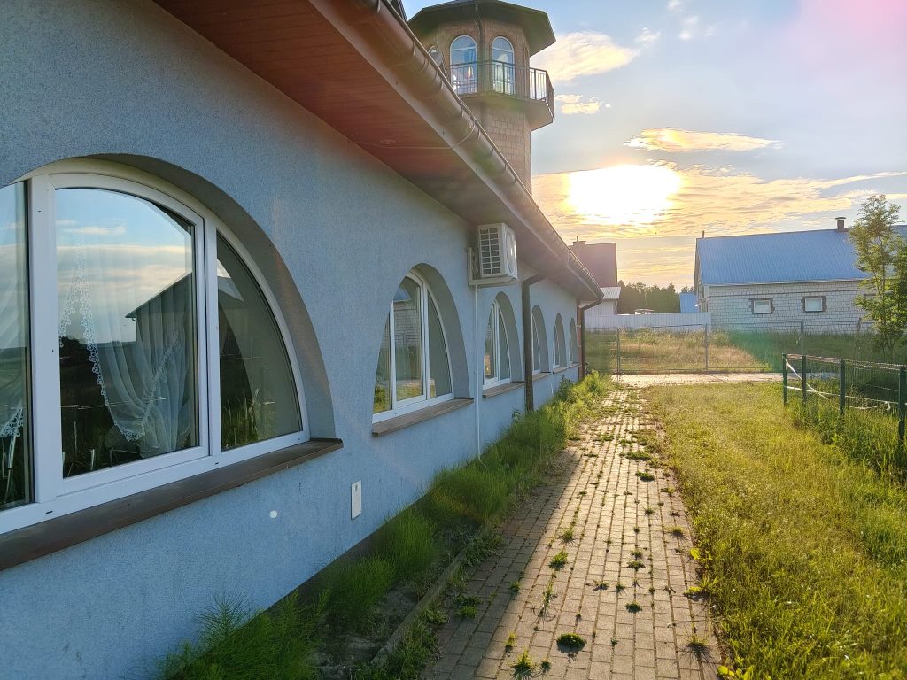 Niebieska ściana budynku z białymi, półokrągłymi oknami, czerwona rynna, wieża z oknami i balkonem, brukowana ścieżka z trawą, drzewa i inne budynki, zachód słońca.
