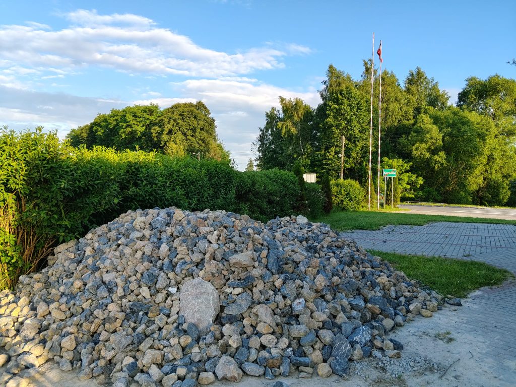 Stos szarych i beżowych kamieni na tle zielonych krzewów, drzew oraz niebieskiego nieba z białymi chmurami, obok żwirowa i brukowana droga.
