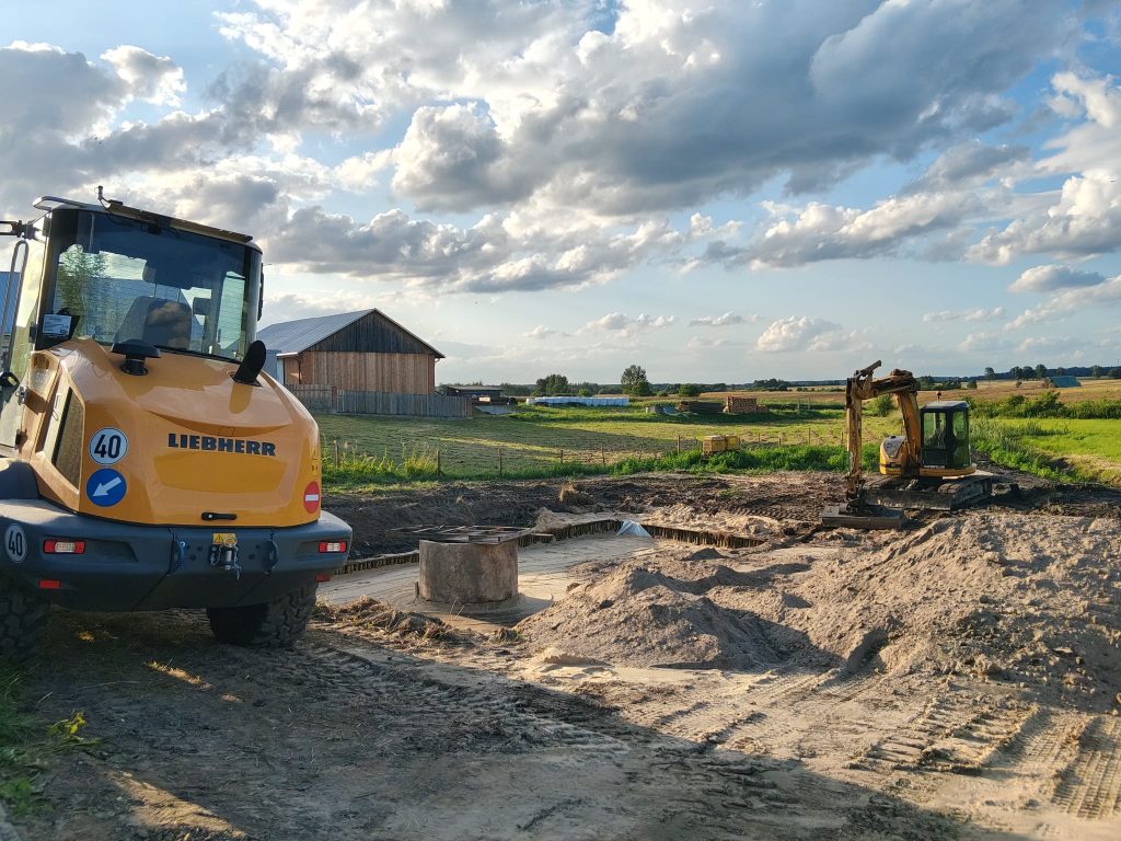 Żółty koparko-ładowarka Liebherr i koparka Caterpillar na tle pola, drewniana stodoła i niebieskie niebo z chmurami.