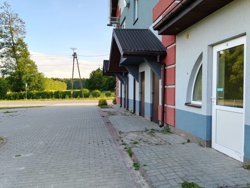 Budynek z niebiesko-czerwonymi ścianami, białymi drzwiami i oknami, daszki nad wejściami, chodnik z szarych kostek, zieleń i drzewa w tle.