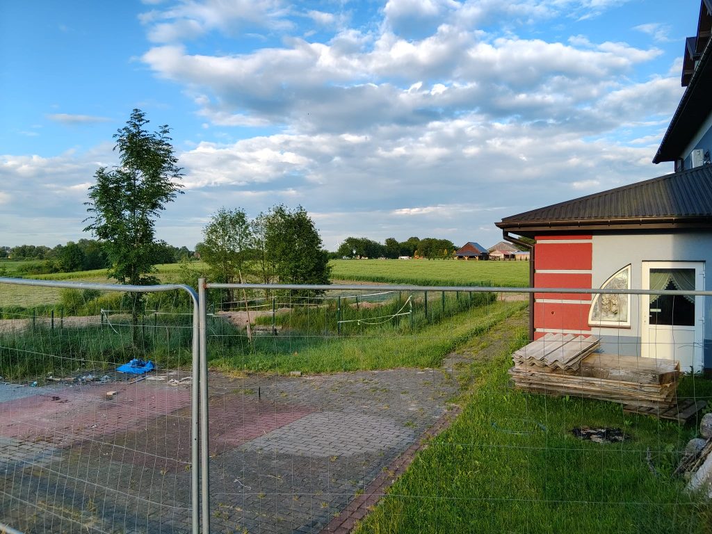 Niska siatka ogrodzeniowa, zielone drzewa i trawa, niebieskie niebo z białymi chmurami, fragment czerwono-szarego budynku z oknem i drzwiami, stos płyt na drewnianej palecie.