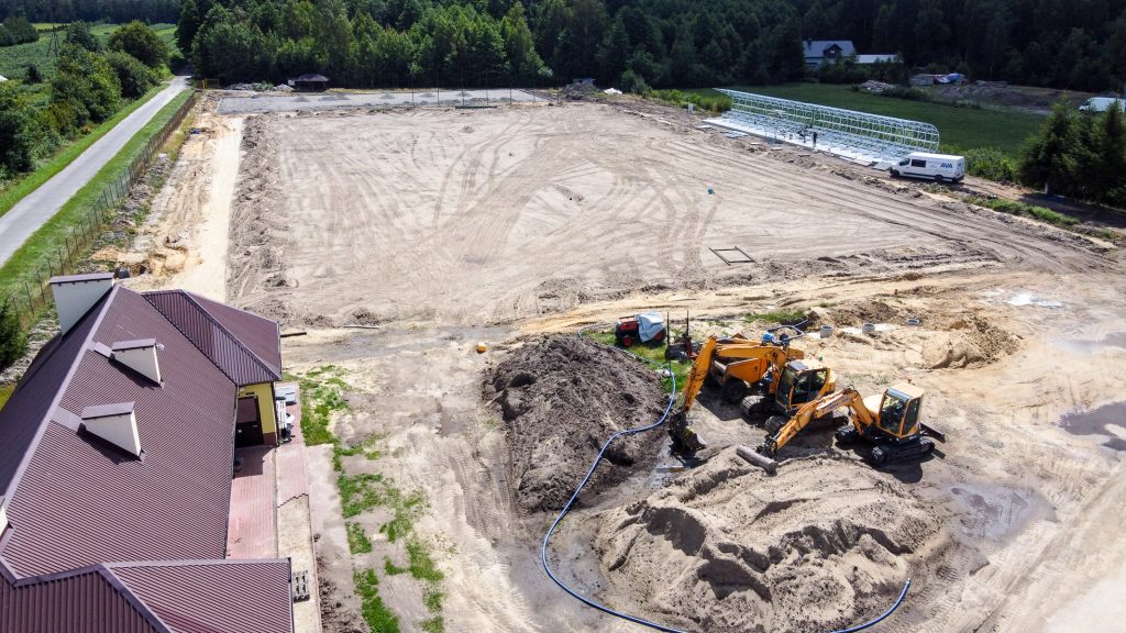Plac budowy z żółtymi koparkami, hałdą ziemi, białą dostawczą furgonetką, częściowo widoczny budynek z brązowym dachem, otoczony zielonymi drzewami i trawnikiem.