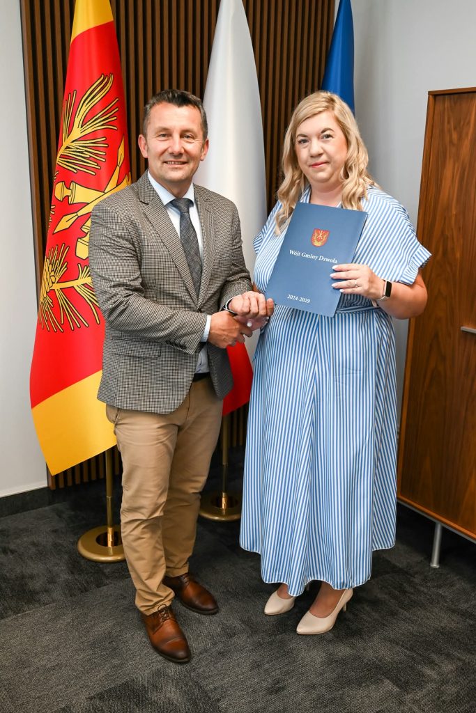 Mężczyzna w jasnej marynarce i beżowych spodniach ściska dłoń kobiety w niebiesko-białej sukience w paski, która trzyma granatową teczkę z napisem "Wójt Gminy Dzwola 2024-2029". W tle trzy flagi.