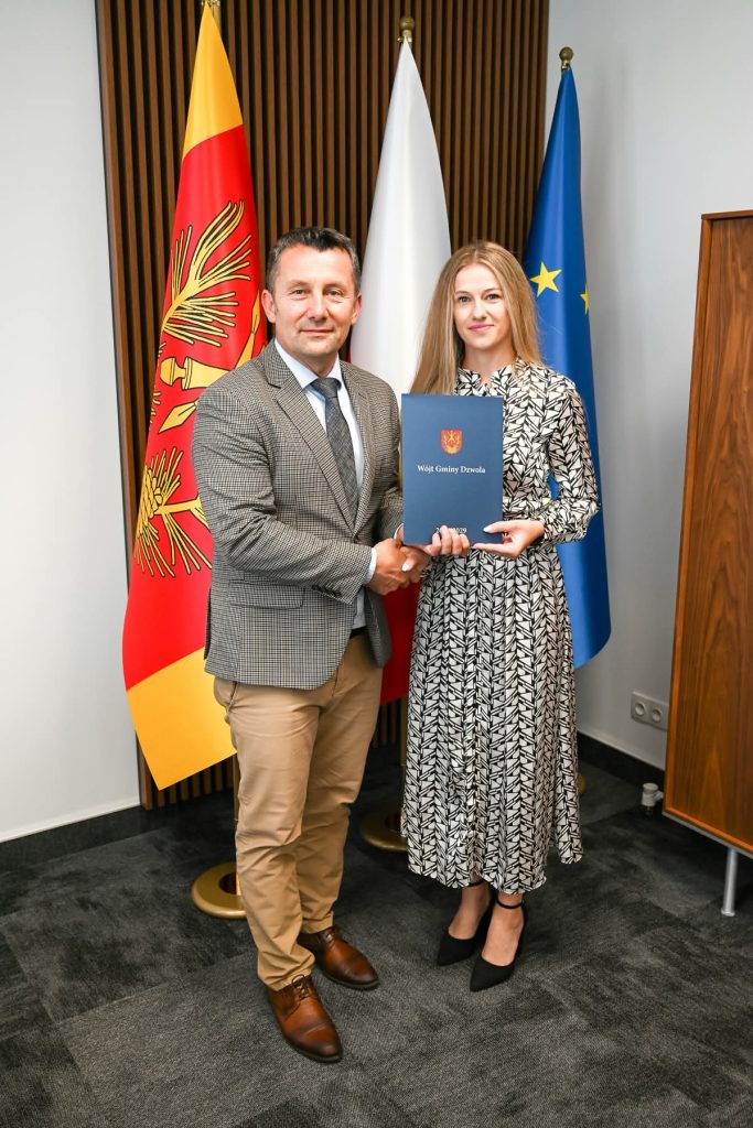 Mężczyzna w beżowych spodniach i marynarce w kratę oraz kobieta w czarno-białej sukience trzymająca granatową teczkę, stoją na tle trzech flag.