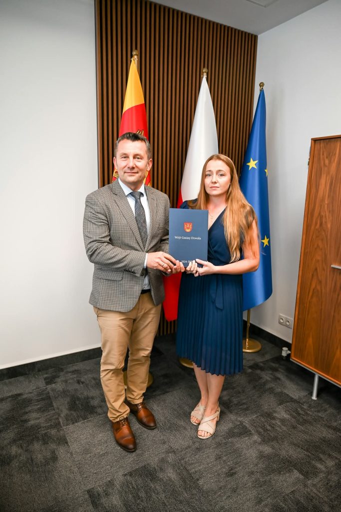 Mężczyzna w marynarce w kratę i beżowych spodniach oraz kobieta w granatowej sukience trzymają niebieski dokument, za nimi flagi i drewniana ściana.