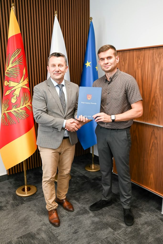 Dwaj mężczyźni ściskają sobie dłonie, jeden trzyma niebieski dokument, w tle trzy flagi: czerwona z żółtym i białą oraz niebieska z gwiazdami.