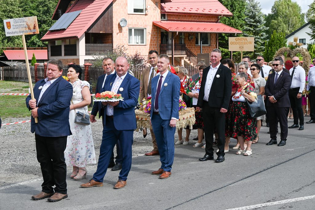 Grupa ludzi ubranych w garnitury i tradycyjne stroje ludowe stoi na ulicy, mężczyzna trzyma tabliczkę z napisem, kobiety z kwiatami i wieńcami, domy i drzewa w tle.