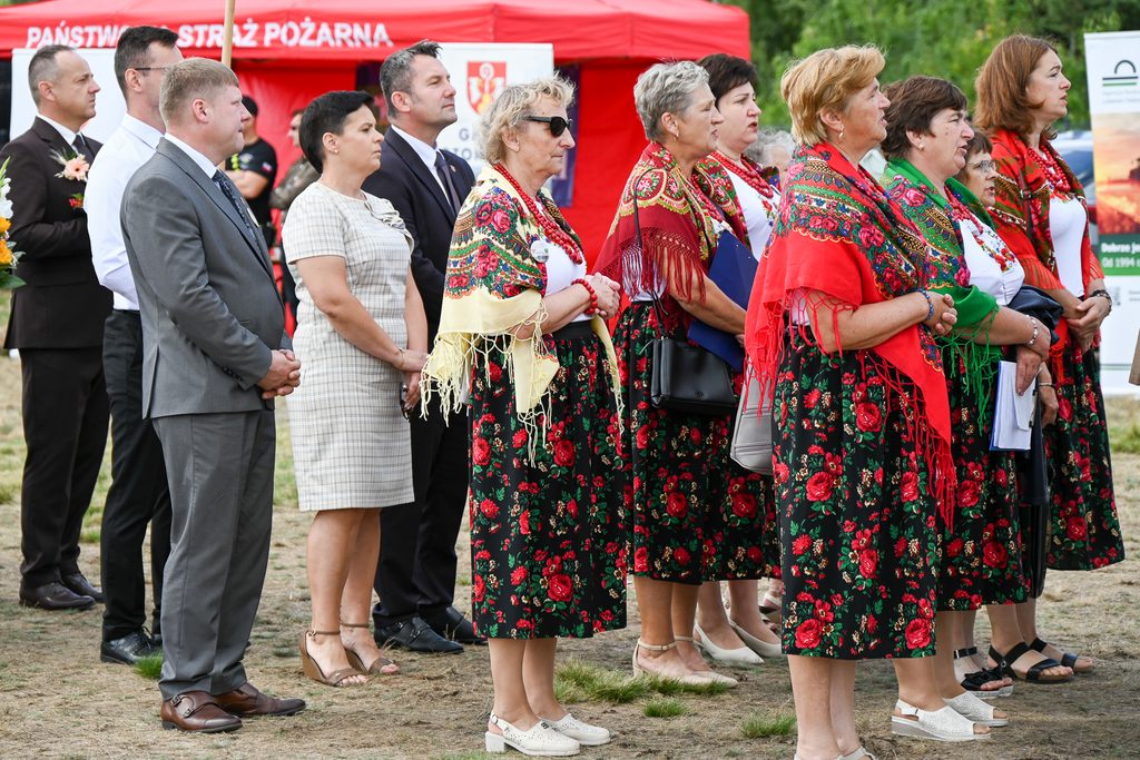 Grupa ludzi stoi na trawie, kobiety w kwiecistych spódnicach i kolorowych chustach, mężczyźni w garniturach, w tle czerwony namiot z napisem Państwowa Straż Pożarna.