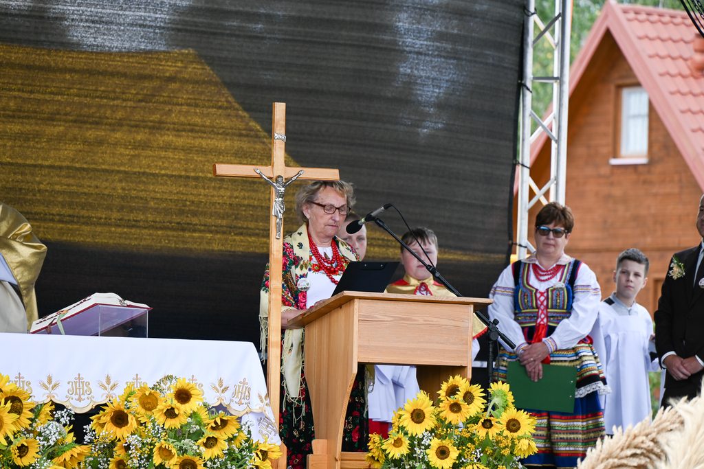 Starsza kobieta w tradycyjnym stroju ludowym przy drewnianym pulpicie z krzyżem, obok dzieci i dorosła kobieta w kolorowym stroju ludowym, w tle drewniany dom i słoneczniki.
