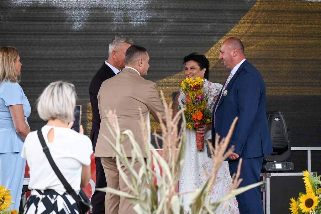 Grupa osób na scenie, kobieta w białej sukni z kwiatowym wzorem trzyma bukiet kolorowych kwiatów, mężczyźni w garniturach, pierwszoplanowo rośliny i kobieta z krótkimi jasnymi włosami z aparatem.