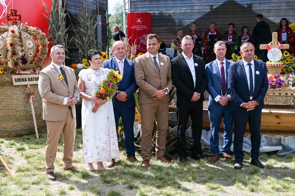 Siedem osób w eleganckich garniturach i biała kobieta w sukience z kwiatowym wzorem, stojąca na zielonej trawie, z wieńcami zbożowymi i kwiatami w tle.