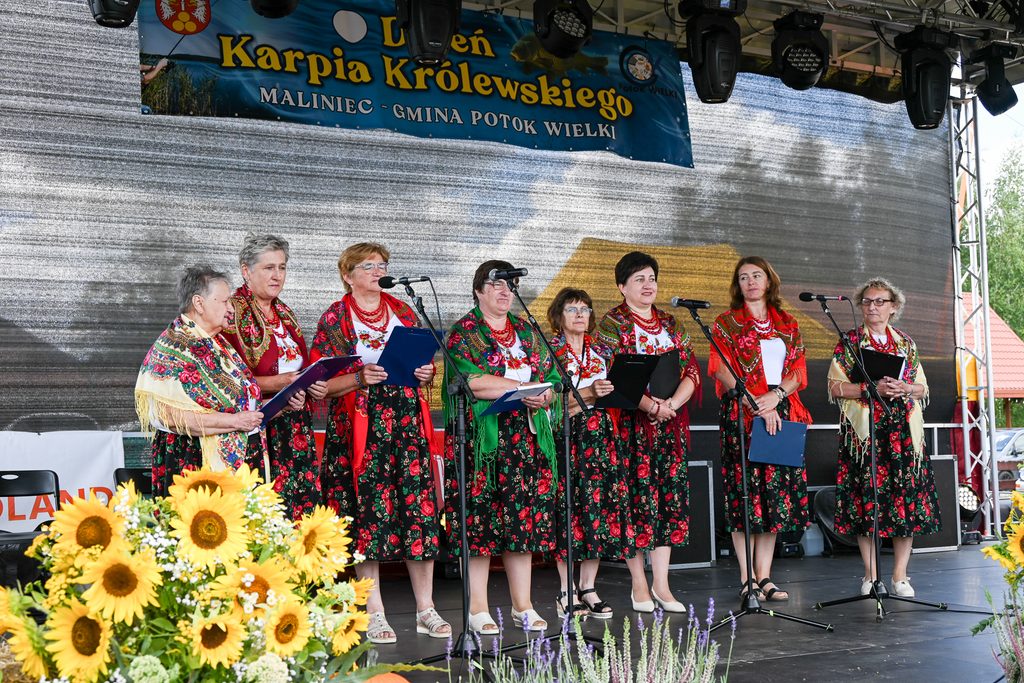 Grupa kobiet w tradycyjnych strojach ludowych z kwiatowymi wzorami i kolorowymi chustami, stojących na scenie z mikrofonami, z napisem i słonecznikami na pierwszym planie.